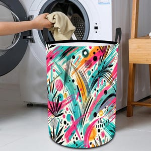 May include: A colorful laundry hamper with a white background and abstract brushstrokes in pink, blue, yellow, and black. The hamper has a black rim and black handles.