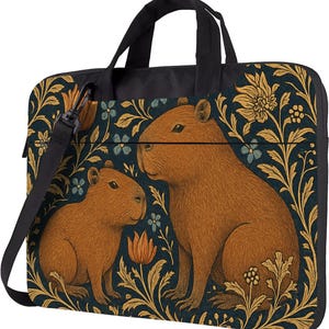 May include: A black laptop bag featuring a brown capybara illustration. The bag has a dark blue background with gold floral and leaf patterns, a black handle, and a detachable shoulder strap.