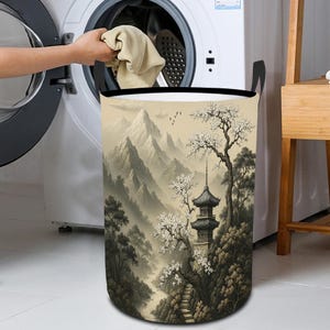 May include: A laundry hamper with a scenic landscape design featuring a pagoda, mountains, and trees in shades of beige, brown, and green. The hamper has black handles and a black rim. A person is putting clothes into a washing machine.