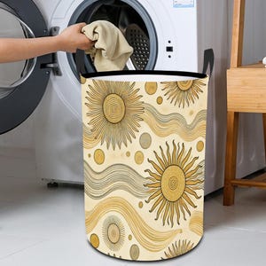 May include: A beige laundry hamper with black handles and a pattern of gold suns and waves. The hamper is sitting in front of a white washing machine.