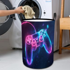 May include: A cylindrical laundry basket featuring a neon-style graphic of a video game controller. The controller design has pink and blue glowing outlines against a dark blue starry background. The basket has black handles.