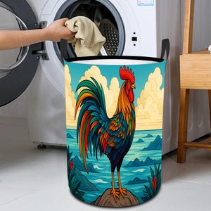 May include: A laundry hamper with a colorful rooster illustration. The rooster is depicted with vibrant blue, green, orange, and red feathers, standing on a rock against a backdrop of water and sky. The hamper is cylindrical with black handles.