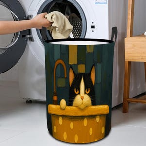 May include: A cylindrical laundry hamper with a whimsical cat design. The hamper features a black and white cat peeking over a yellow dotted rim. The background has a dark teal and yellow artistic design. Black handles are on the sides.