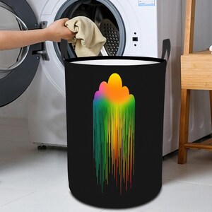 May include: A black laundry basket with a vibrant, abstract design. The design features a cloud-like shape at the top that transitions into vertical lines of rainbow colours. The basket has black handles and a white interior.