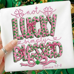 Puede incluir: Camiseta blanca con texto bordado: "not LUCKY simply BLESSED" en rosa y verde. La palabra "LUCKY" está llena de tréboles verdes. El texto "Romans 4:7-8" está en la parte inferior. La camiseta está sujeta por una mano.