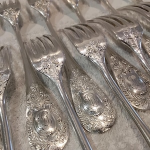 Pode incluir: Uma coleção de garfos de metal prateado com padrões florais intrincados em relevo nos cabos. Cada cabo exibe um cartucho central em forma de escudo gravado com um monograma decorativo. Os garfos possuem quatro dentes e acabamento polido.