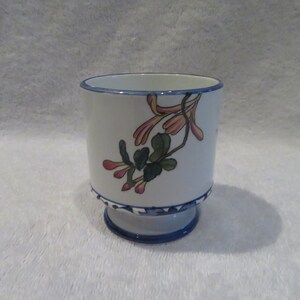 May include: A white ceramic cup with a blue rim and a floral design. The design features pink and yellow flowers with green leaves.