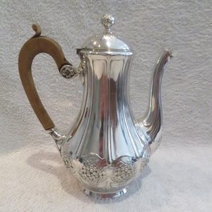 Pode incluir: Uma cafeteira de prata com uma pega de madeira e um design floral. A cafeteira tem um acabamento brilhante e um corpo redondo.