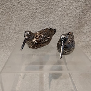 Op de afbeelding: Twee gedetailleerde, zilverkleurige vogelbeeldjes met lange snavels en een getextureerd lichaam. Ze staan op een helder, schuin oppervlak. De beeldjes zijn waarschijnlijk decoratieve items, voor een huis of kantoor.