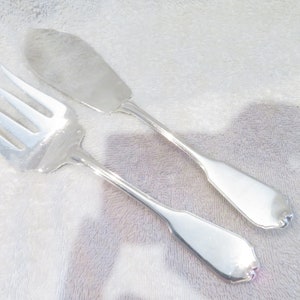 May include: A silver-colored fork and butter knife set. The fork has three tines, and the butter knife has a wide, flat blade. Both utensils have a decorative, scalloped edge on the handle.