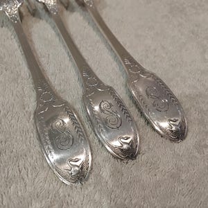 May include: Three silver-toned metal spoons featuring an engraved letter S in the center of the handle. The handle ends are decorated with a detailed relief of a swan with spread wings, surrounded by an engraved leaf pattern along the edges.