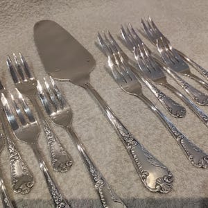 Pode incluir: Uma coleção de garfos de servir e uma espátula para bolo prateados. Os talheres apresentam detalhes florais ornamentados nas alças. As peças estão dispostas sobre uma superfície clara e texturizada.