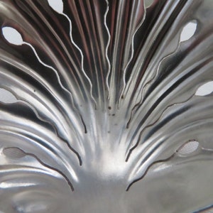 May include: A silver metal tea strainer with a decorative, fan-shaped design. The strainer has a series of small holes and a scalloped edge.
