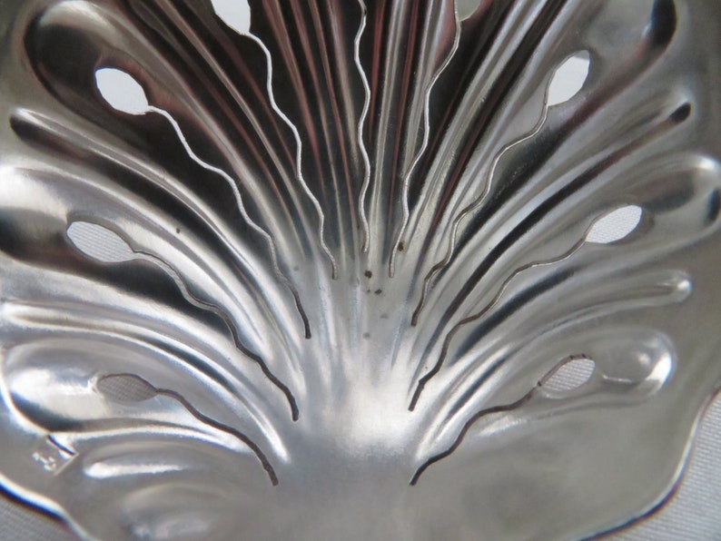 May include: A silver metal tea strainer with a decorative, fan-shaped design. The strainer has a series of small holes and a scalloped edge.