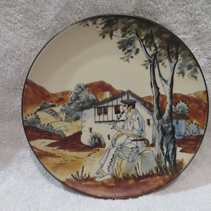 May include: A ceramic plate with a hand-painted scene of a person playing a flute in front of a white house with a brown roof. The scene is set in a rural landscape with trees and hills. The plate has a brown rim.