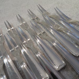 Op de afbeelding: Een verzameling zilverkleurige vorken met drie tanden. De vorken hebben een slanke, gepolijste uitstraling, met een licht gebogen handvat en een decoratief patroon aan de basis. De tanden zijn scherp en gelijkmatig verdeeld.