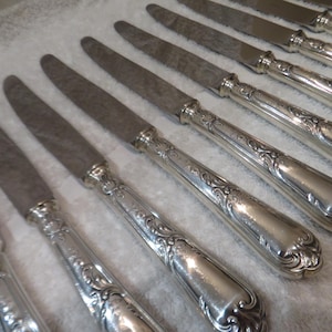Puede incluir: Un juego de cuchillos de cena de plata con mangos adornados. Los cuchillos están dispuestos en una fila sobre una superficie blanca.