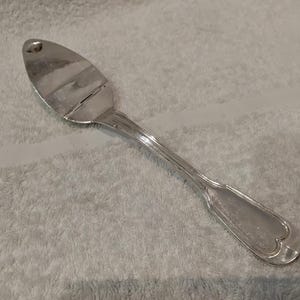 Peut inclure: Un ustensile de service en métal argenté doté d'une tête large, plate et ovale, ainsi que d'un manche fin et décoratif. Le manche présente un motif strié subtil et une extrémité arrondie en forme de cœur. La surface du métal est polie et brillante.