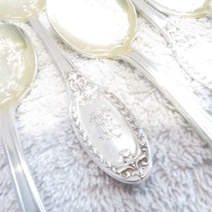May include: A set of silver spoons with ornate handles. The spoons are laid out on a white fluffy towel. The handle of one spoon has an engraved monogram.