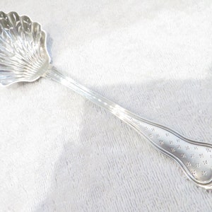 Peut inclure: Une cuillère de service en argent avec une cuillère en forme de coquillage et un long manche décoré. Le bol a un design détaillé en forme d'éventail. La poignée présente un motif délicat de petites formes florales. La cuillère est probablement utilisée pour servir du sucre.