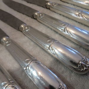 Könnte beinhalten: Ein Set aus sechs versilberten Essmessern mit verzierten Griffen. Die Messer haben eine polierte Oberfläche und sind in einer Reihe angeordnet.