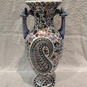 Peut inclure: Un vase en céramique de style amphore avec une base blanche ornée d'un motif floral et cachemire dense et multicolore dans des tons de bleu, rouge et vert. Le vase possède deux anses bleues et une petite étiquette rectangulaire Fabrika D'Art.