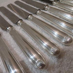 May include: A set of silver-plated dinner knives with ornate handles. The handles have a shell-like design at the end.