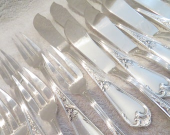 May include: A set of silver forks and knives with ornate handles. The silverware is arranged in a row on a white surface.