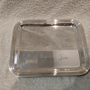 Può includere: Un vassoio da portata rettangolare color argento con una superficie lucida e riflettente. Il vassoio presenta un bordo rialzato a più livelli e diverse firme o iscrizioni scritte a mano incise al centro della base metallica.