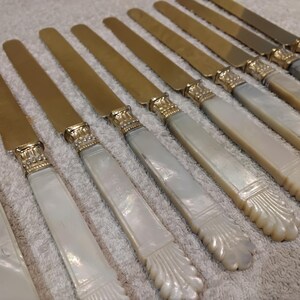 May include: A set of antique butter knives with gold-colored blades and ornate, pearlescent handles. The handles have a decorative, carved design at the base. The knives are arranged diagonally on a white surface.