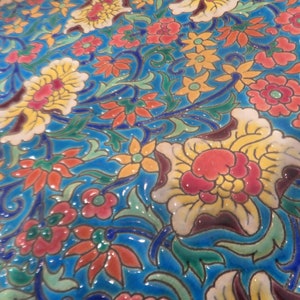 Pode incluir: Um close-up de um azulejo de cerâmica azul com um padrão floral colorido. O azulejo apresenta uma variedade de flores, incluindo flores vermelhas, amarelas e brancas, com folhas e videiras verdes. O azulejo tem um acabamento brilhante e um design em relevo.