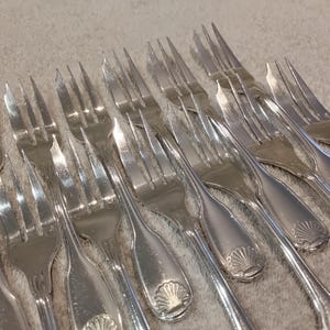 Puede incluir: Un conjunto de tenedores plateados con tres púas, dispuestos en diagonal. Los tenedores tienen una superficie brillante y reflectante y un diseño decorativo de concha en el mango. El fondo es una superficie clara y texturizada.