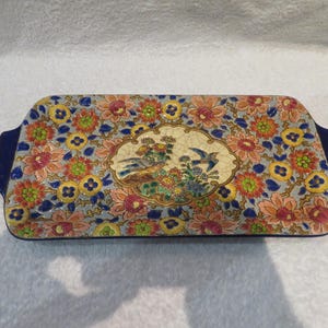 Peut inclure: Plateau de service rectangulaire en céramique avec un motif floral dans les tons orange, rose, jaune et bleu. Le plateau a un motif ovale central avec des oiseaux et des fleurs. Les poignées sont de couleur bleu foncé.