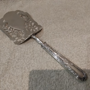May include: Silver-colored cake server with an ornate, openwork design on the blade and handle. The blade has a decorative, scalloped edge and a central, solid oval. The handle is detailed with floral patterns.