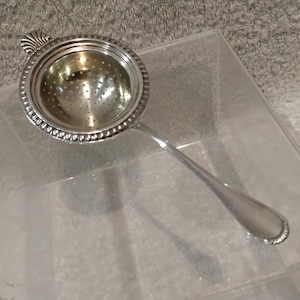 Op de afbeelding: Een theezeef van zilverkleurig metaal met een ronde kom met geperforeerde bodem, een decoratieve kralenrand en een schelpvormige handvatbevestiging. De zeef heeft een lang, glad en taps toelopend handvat aan de zijkant van de kom.