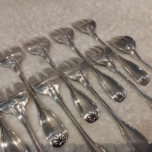 May include: A collection of silver serving forks with three prongs. The forks have a decorative floral design at the base of the handle. The forks are arranged on a light-coloured surface.