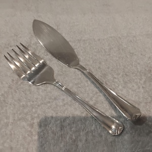 May include: A silver fork and butter knife. The fork has four tines and a long handle. The butter knife has a rounded blade and a matching handle. The cutlery is placed on a grey surface.