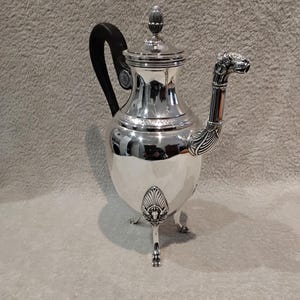 May include: A silver coffee pot with a black handle and a decorative animal head spout. The pot has a lid with a finial and stands on three ornate feet. The pot is highly reflective, with a polished finish.