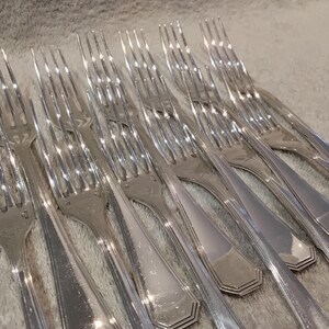 Pode incluir: Uma coleção de garfos prateados dispostos sobre uma superfície cinza claro. Os garfos têm dentes e um design de cabo decorativo. Os talheres são reflexivos, sugerindo um acabamento polido, e são adequados para jantares formais.
