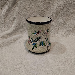 Peut inclure: Un mug en céramique avec une finition émaillée craquelée de couleur crème et un bord foncé. L'extérieur est orné d'un motif peint à la main représentant un scarabée vert sur une branche avec des fleurs roses et des feuilles vertes, forme cylindrique.