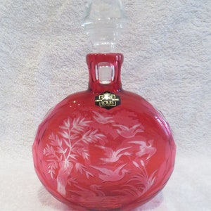 Puede incluir: Una decantador de vidrio rojo con un tapón de vidrio transparente. La decantador tiene un cuerpo redondo con un diseño grabado en blanco de pájaros y árboles. La decantador está etiquetada "Saint Louis Crystal."