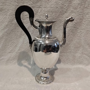 May include: A silver coffee pot with a black handle. The pot features a rounded body, a decorative spout, and a lid with a small finial. The handle is curved and the spout is shaped like a dragon's head. It stands on a pedestal base.