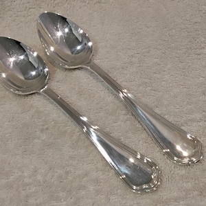 Può includere: Due cucchiai d'argento lucido con superficie riflettente su uno sfondo grigio chiaro strutturato. I cucchiai hanno un design classico con un bordo decorativo sul manico e una coppetta arrotondata.