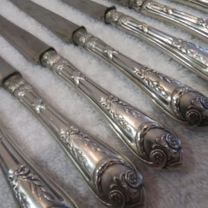 May include: A set of six silver-plated dinner knives with ornate handles. The handles feature a floral design and are slightly curved. The blades are sharp and pointed.