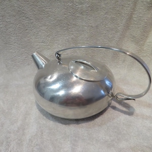 Peut inclure: Une théière argentée avec un corps arrondi, une anse incurvée et un couvercle avec une petite poignée. Le bec est incliné et la théière repose sur une surface de couleur neutre. La théière est en métal.