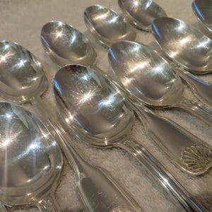 May include: A collection of shiny, silver spoons. The spoons have an oval bowl and a decorative handle with a shell design. The spoons are arranged in a close-up shot, reflecting light.