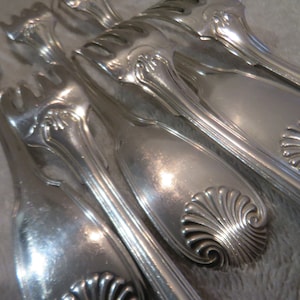 May include: Close-up of several pieces of antique silverware. The silver utensils include serving spoons and forks with ornate, floral-like designs and shell-shaped details. The silverware is arranged on a light-coloured surface.
