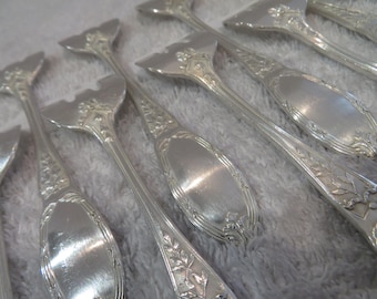 Op de afbeelding: Een set zilveren serveerbestek met sierlijke handvatten en een decoratief ontwerp. De bestekken hebben een gepatineerde afwerking en zijn in een rij gerangschikt.