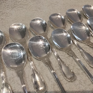 Op de afbeelding: Een verzameling gepolijste zilveren lepels met ovale kommen en lange, slanke handvatten. De lepels zijn in een licht overlappend patroon gerangschikt en reflecteren licht. Het zilverwerk is waarschijnlijk bedoeld om te serveren of te dineren.