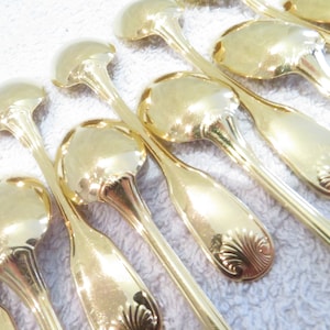 Pode incluir: Um conjunto de colheres banhadas a ouro com cabos ornamentados. As colheres estão dispostas em fila, com os cabos virados para o observador. As colheres são todas do mesmo tamanho e formato, e têm um acabamento brilhante e polido.